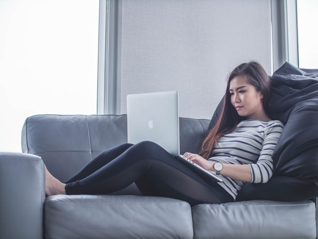 Uma mulher sentada sobre um sofá com um laptop sobre as pernas.